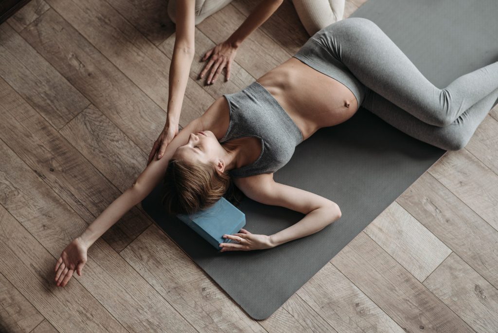 L'image montre une femme enceinte en train de pratiquer le yoga avec un sourire heureux et une posture parfaite. Elle porte une tenue de yoga blanche et rose et est entourée de plantes et de couleurs vives.